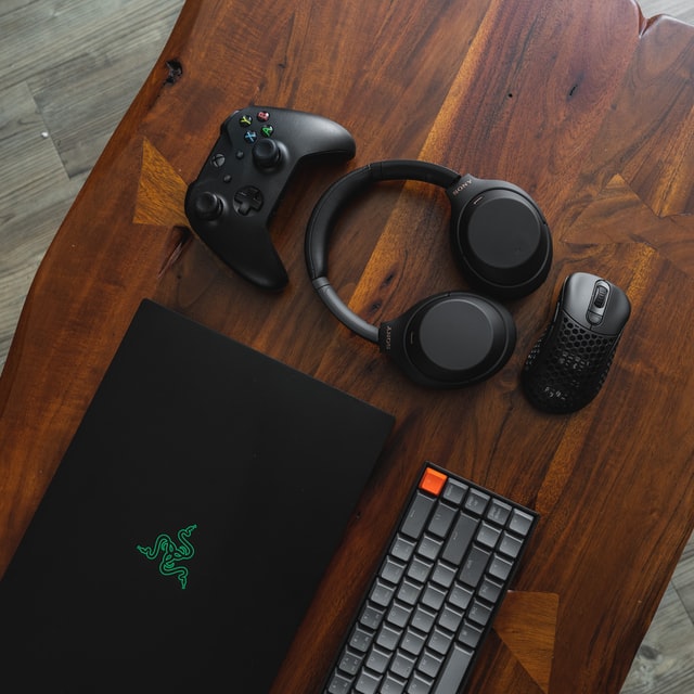 Czarny laptop Razer, szara klawiatura z pomarańczowym przyciskiem, czarna myszka, czarne słuchawki bezprzewodowe nauszne i czarny pad od Xboxa