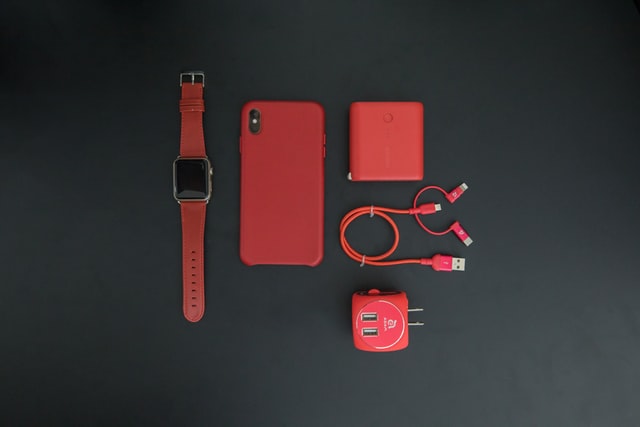 Gadżety i akcesoria w kolorze czerwonym na czarnym tle. Od lewej zegarek, telefon w pokrowcu, słuchawki w etui ładującym, kabel USB, ładowarka z złączem amerykańskim