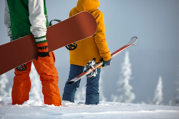 Dwoje ludzi trzyma w rękach narty i deskę snowboardową. W tle śnieżny krajobraz gór.